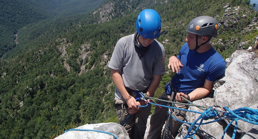 Two people wearing safety gear stand on a rock ledge above a green wooded area. One of them is an instructor, providing direction to the other. 